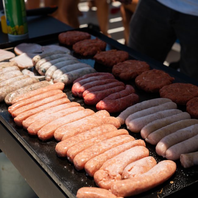 A variety of sausages and hamburger patties are being grilled on an outdoor barbecue, with drinks and blurred figures in the background, creating the ideal ambiance for a Catamaran party Sydney.