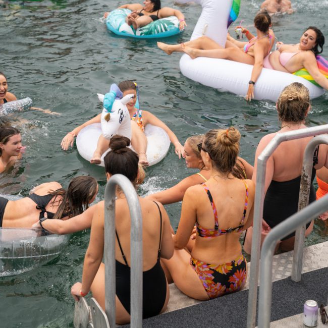 People enjoying a day at the lake, swimming, and lounging on inflatable floats. Some individuals are sitting on the dock while others are in the water, much like a Catamaran party Sydney where everyone revels in relaxation and fun.