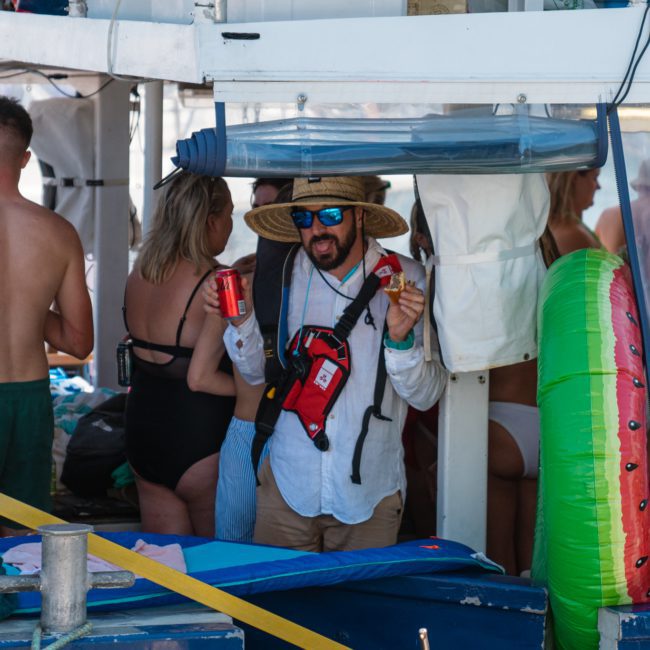 A person wearing a hat and sunglasses holds a drink and gestures at a gathering on a boat. Other people are present, some in swimwear. An assortment of inflatables is visible in the background, highlighting the fun atmosphere of this catamaran party in Sydney.