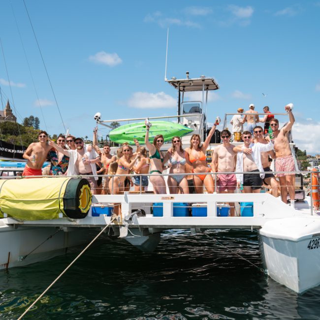 A group of people in swimsuits stand and celebrate on the deck of a white catamaran, surrounded by other boats on a sunny day during a lively Sydney boat party hire.