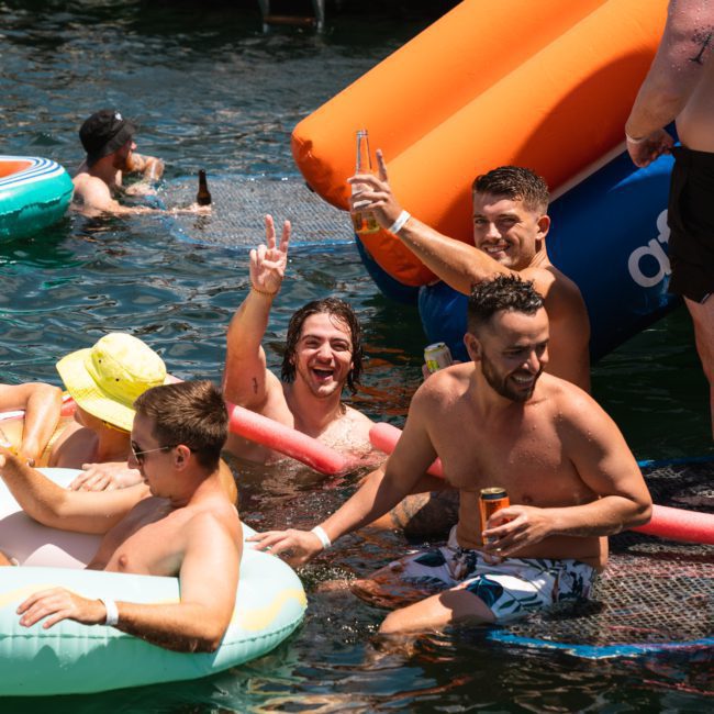 A group of people enjoying a sunny day in the water with inflatable floats and drinks near a luxury yacht. One man is smiling and making a peace sign, embodying the perfect vibe of a catamaran party in Sydney.