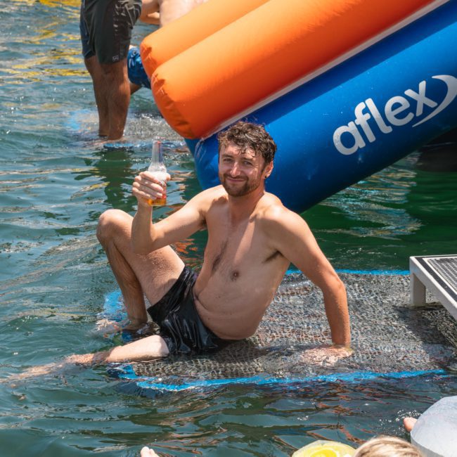 A man with a beard sits partially submerged in water, holding a drink in his right hand, in front of an inflatable structure labeled "aflex." Around him, other individuals enjoy the water as part of a luxury yacht hire on Sydney Harbour.