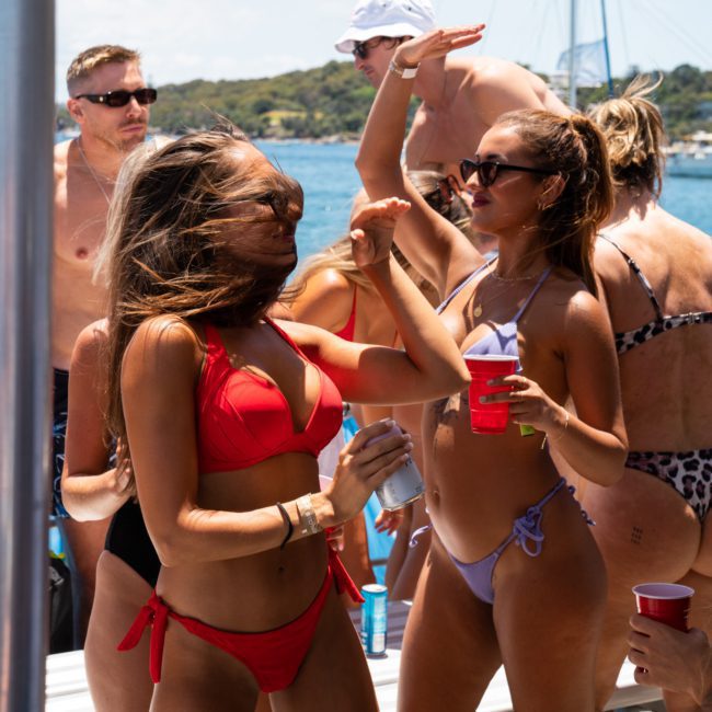 A group of people in swimwear dance and interact on a luxury yacht under sunny weather. Several of them are holding red cups, and a marina is visible in the background.