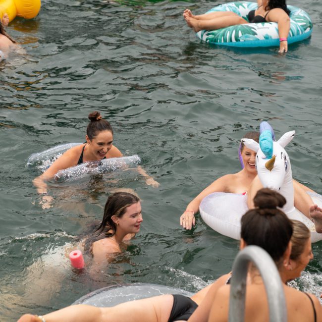 Several people are enjoying a swim in a body of water, some using various inflatable pool floats, including a unicorn-shaped one. Nearby, a luxury yacht hire Sydney adds an extra touch of elegance to their day on the sparkling waters.