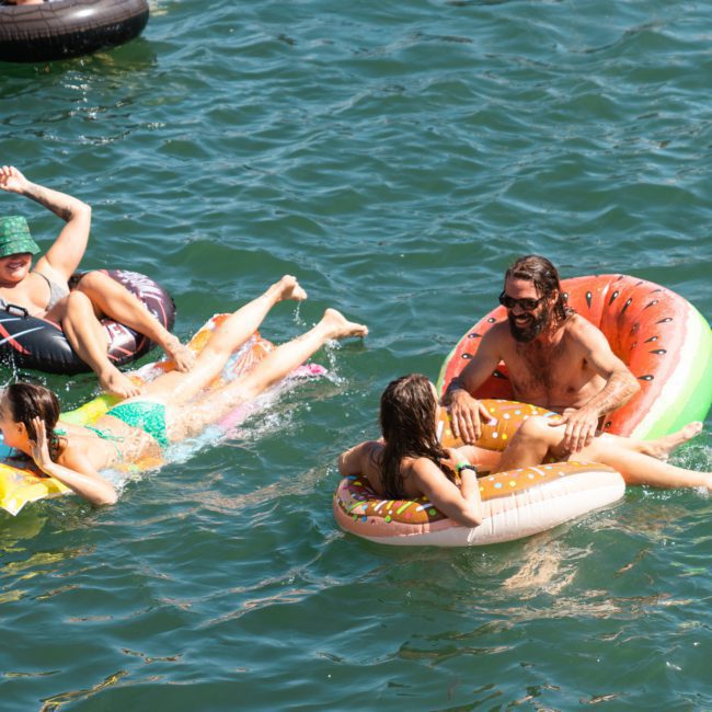 Four people floating on colorful inflatables in a body of water. One person is on a doughnut-shaped float, another on a watermelon-shaped float, and two are on patterned round floats. Enjoy the ultimate relaxation or amp up the fun with a DJ boat hire Sydney for an unforgettable experience.