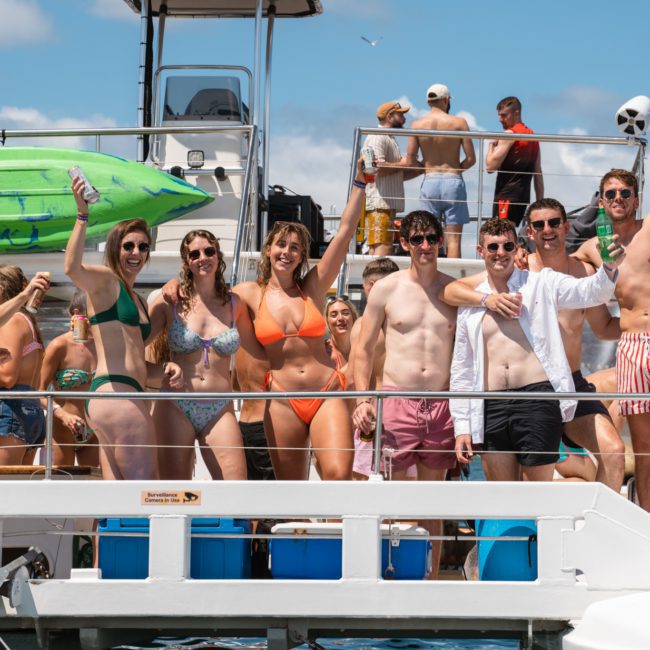 A group of people in swimwear stand and smile on a private yacht charter in Sydney Harbour under a sunny sky, with others and watercraft in the background.