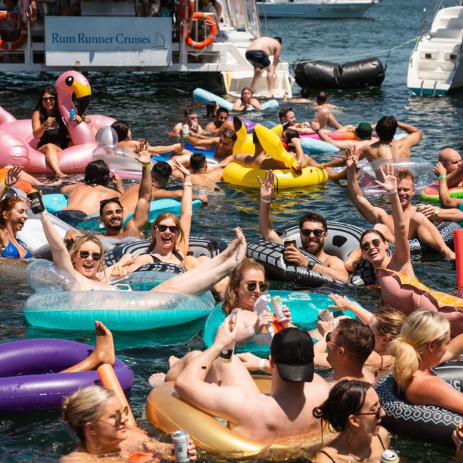 A vibrant group of people enjoying a sunny day on a lake, floating on various inflatable devices near a boat. Some are posing for the camera, while others relax and converse in the water, capturing the essence of a memorable Sydney boat party hire.