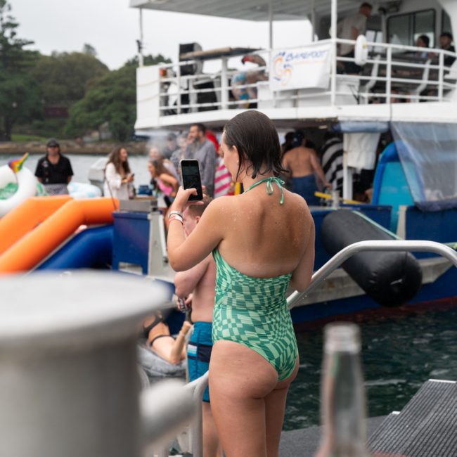A woman in a green swimsuit stands on a dock, holding a phone. People are gathered on a nearby catamaran with inflatables in the water, suggesting a lively party by the water.