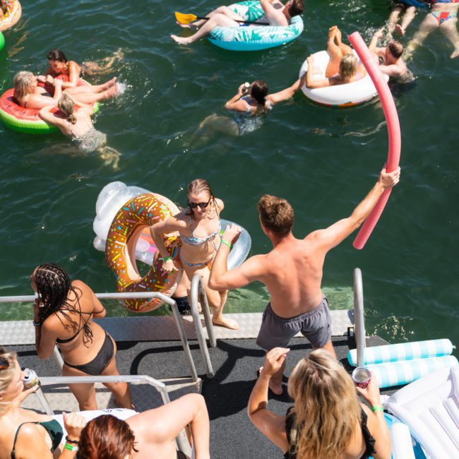 A group of people enjoying a sunny day at a lake, using various inflatable pool toys and floating devices. Some are entering the water from a dock while others are swimming and playing. The scene could easily rival the fun of a luxury yacht hire in Sydney Harbour.