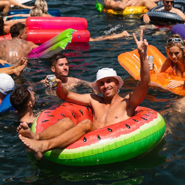 A group of people are floating on inflatable pool toys in the water. One man in a watermelon float holds a drink and flashes a peace sign, making it feel like a mini Sydney boat party hire event.