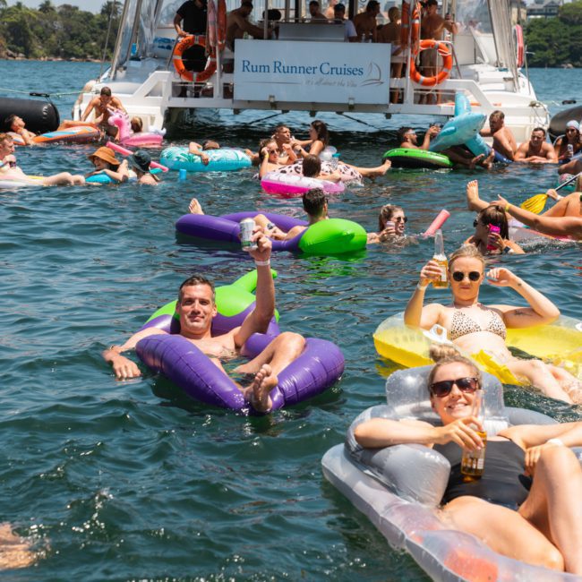 A group of people relaxes and floats on inflatable loungers in the water near a boat labeled "Rum Runner Cruises" on a sunny day, enjoying a Sydney boat party hire.