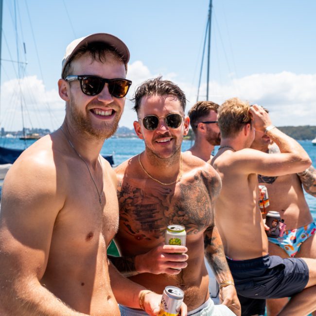 A group of young men enjoying drinks on a sunlit private yacht charter in Sydney Harbour, with other boats and a bridge visible in the background.