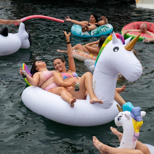 People are floating on inflatable pool toys in a body of water, including a large unicorn float. Two women in swimsuits are sitting on the unicorn float while others relax nearby, enjoying the vibes reminiscent of a private yacht charter Sydney Harbour.