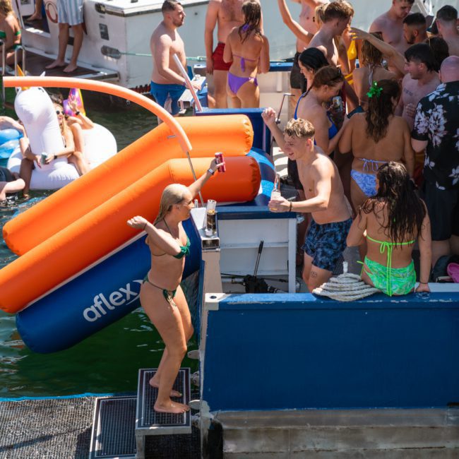 A group of people are enjoying a party on a private yacht charter in Sydney Harbour, some are dancing and others are on large inflatable pool floats in the water.