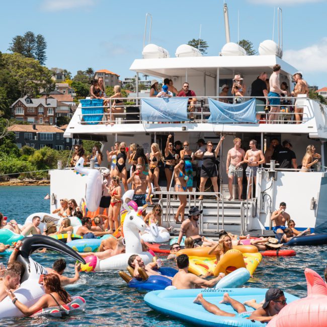 A large group of people enjoys a sunny day on a yacht and in the water using various inflatables, with a scenic coastal town in the background, perfect for a Catamaran party Sydney.