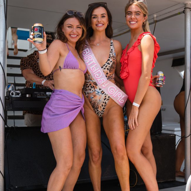 Three women in swimsuits, one wearing a "Birthday Girl" sash, holding drinks and posing on a private yacht charter Sydney Harbour.