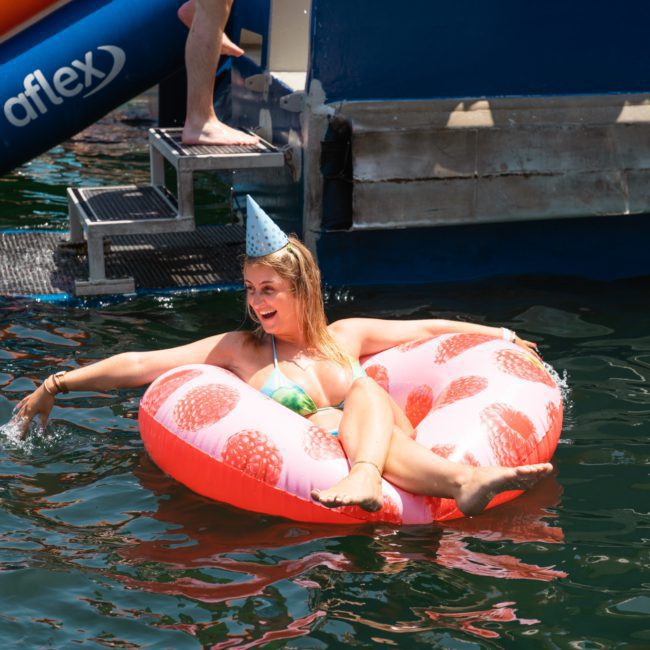 A person wearing a party hat is sitting on a pink inflatable tube with a red heart pattern, floating in the water near a blue structure. In the background, you can see a private yacht charter in Sydney Harbour, adding to the festive ambiance.