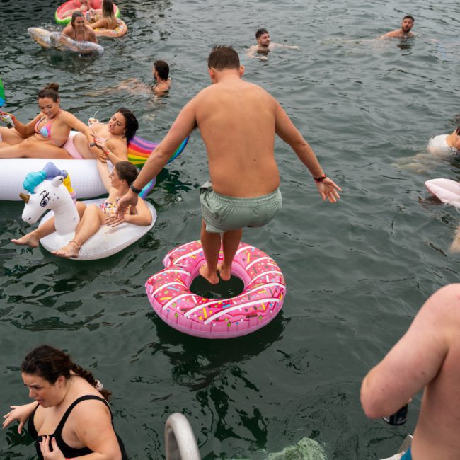 A group of people enjoying a pool party with various inflatable toys. One person stands on a pink donut floatie in the middle of the water, reminiscent of the fun and elegance found at Catamaran party Sydney events.