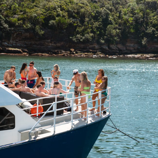 A group of people socialize on the deck of a luxury yacht hire Sydney anchored near a forested coastline under clear skies.