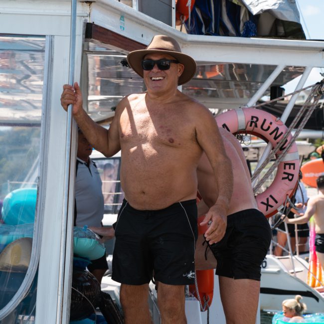 A shirtless man in a hat and sunglasses stands on a boat, smiling. Another person wearing swim trunks is nearby, partially obscured. Rings and equipment are visible around the boat, suggesting a fun time during a private yacht charter Sydney Harbour.