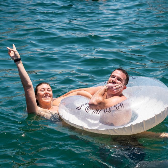 Two people float in clear blue water; one holds up a peace sign while the other rests his head on an inner tube labeled "BARDAK," enjoying what feels like a private yacht charter in Sydney Harbour.
