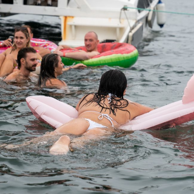 A group of people swim and float in the sea near a boat, using various inflatable rafts, including a flamingo-shaped float, as part of a lively catamaran party Sydney experience.