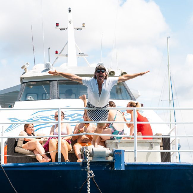A group of people relax on the deck of a boat under a partly cloudy sky. A man stands at the front with his arms outstretched, while others sit in the background, enjoying a luxurious yacht hire in Sydney.