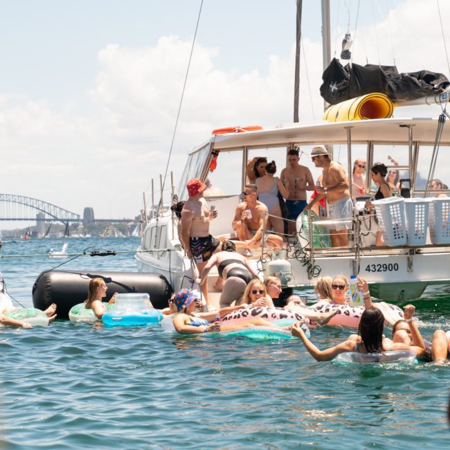 People relaxing on inflatable floats and swimming near anchored boats in a sunny, crowded waterway with a bridge visible in the background, possibly enjoying a Catamaran party Sydney event.