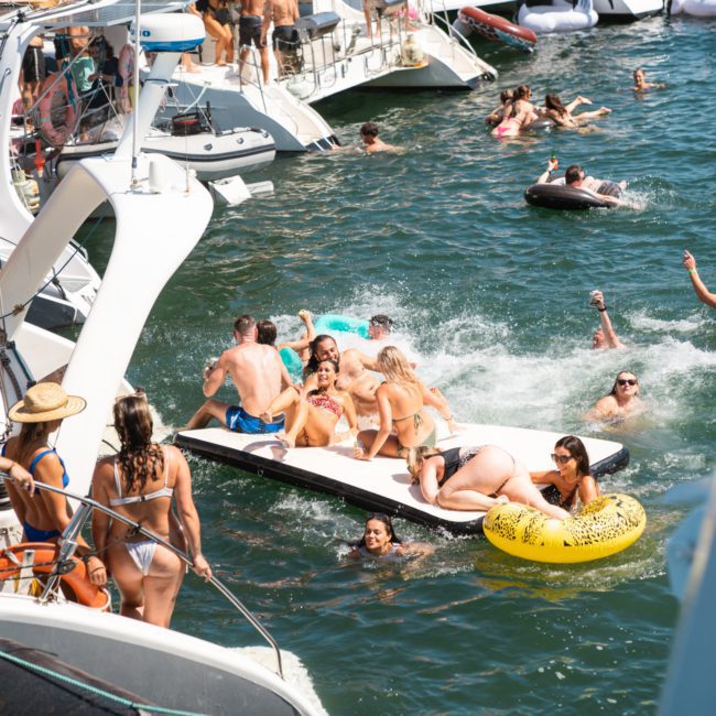 Groups of people in swimwear are enjoying a sunny day on the water, gathered around boats and floating devices, with some swimming and others lounging. Experience the ultimate catamaran party Sydney has to offer, perfect for a memorable day with friends or colleagues.