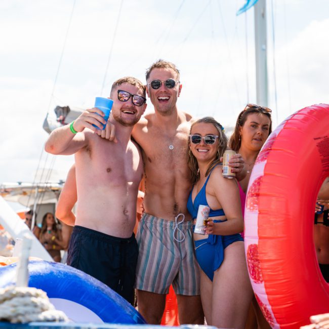 Group of people in swimwear enjoying a sunny day on a luxury yacht hire in Sydney. One person is holding a red inflatable ring, and others are holding drinks.