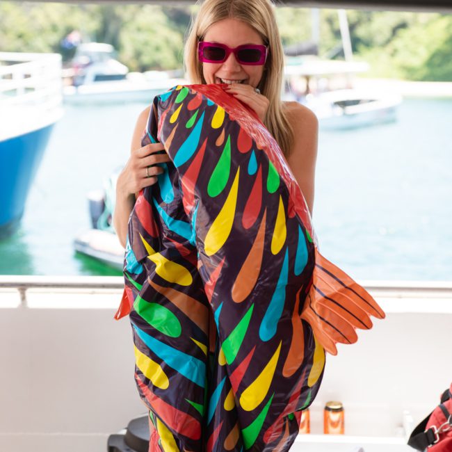 A person wearing pink sunglasses and a swimsuit holds an inflatable fish on a boat with water and other boats in the background, perfect for a Sydney boat party hire.
