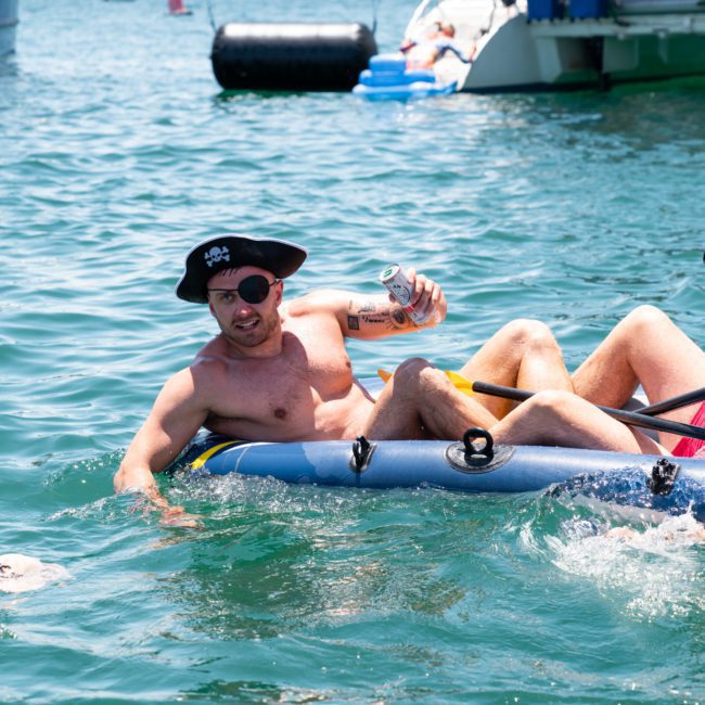 Two men in swim trunks are in a small inflatable boat on the water. One is wearing a pirate hat and holding a beverage, while the other paddles. In the background, a luxurious private yacht charter Sydney Harbour awaits.