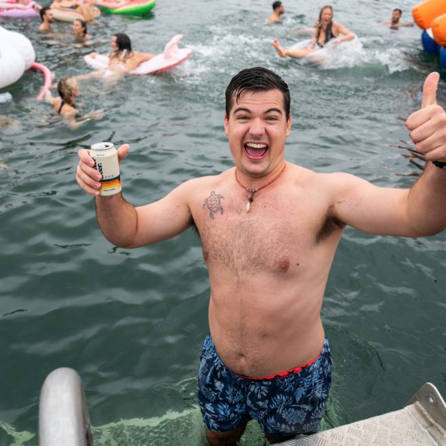 A man in swim trunks, holding a can, stands in the water near a boat hired for a catamaran party Sydney, smiling and giving a thumbs-up, while others swim and float behind him.