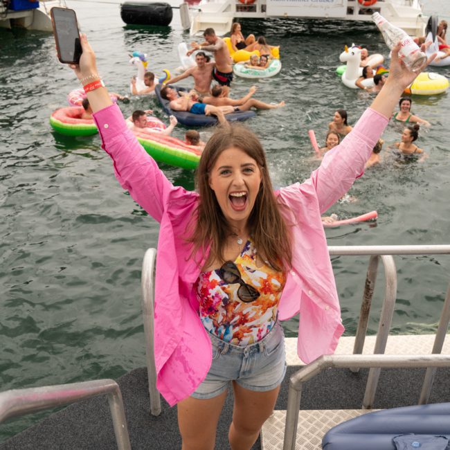 A woman in a pink shirt cheers with raised arms on a boat as people swim and float nearby, enjoying a Sydney boat party hire. A group is on another boat in the background.