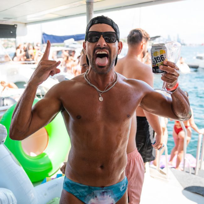 A man wearing a shark-themed swimsuit and sunglasses holds a can of beer while making a rock-on gesture at a private yacht charter on Sydney Harbour.