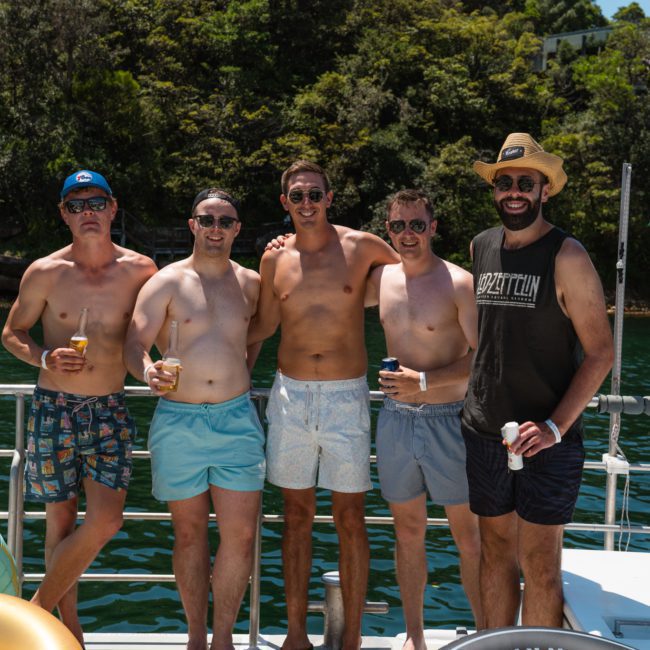 Five men in swimwear and sunglasses stand together on a boat, holding drinks. They are surrounded by inflatable pool toys, with a backdrop of water and greenery, perfect for a catamaran party in Sydney.