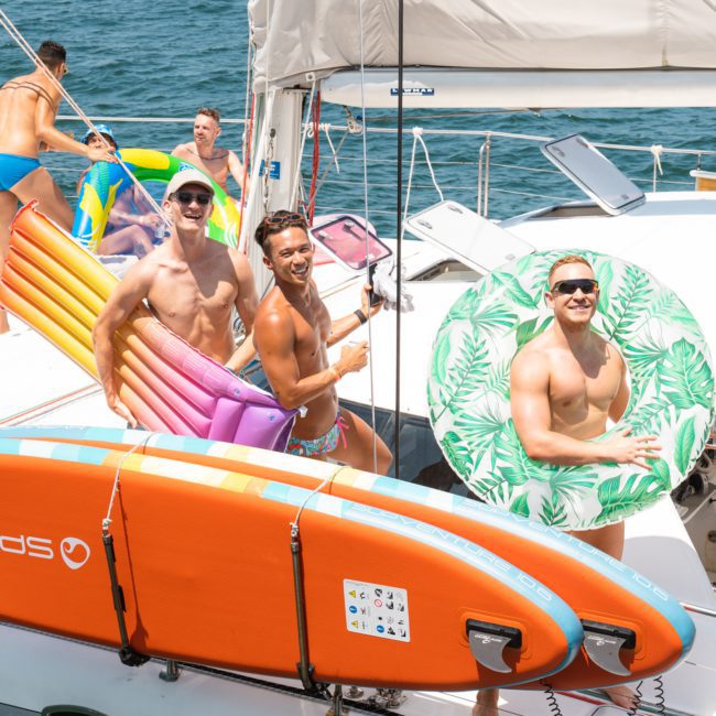 Four men are on a boat, smiling at the camera. They are holding inflatable pool floats and a paddleboard. The boat is on the water under sunny skies—a perfect day for Sydney boat party hire or luxury yacht hire Sydney.