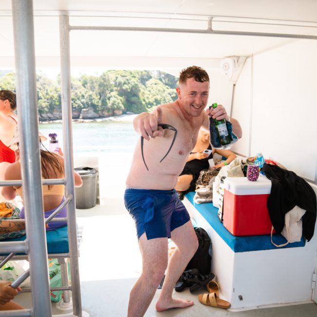 A man in swim shorts holds snorkel gear and a drink while standing on a boat's deck. Other people and personal items are visible in the background. The scene overlooks water and greenery, perfect for a catamaran party Sydney enthusiasts will love.