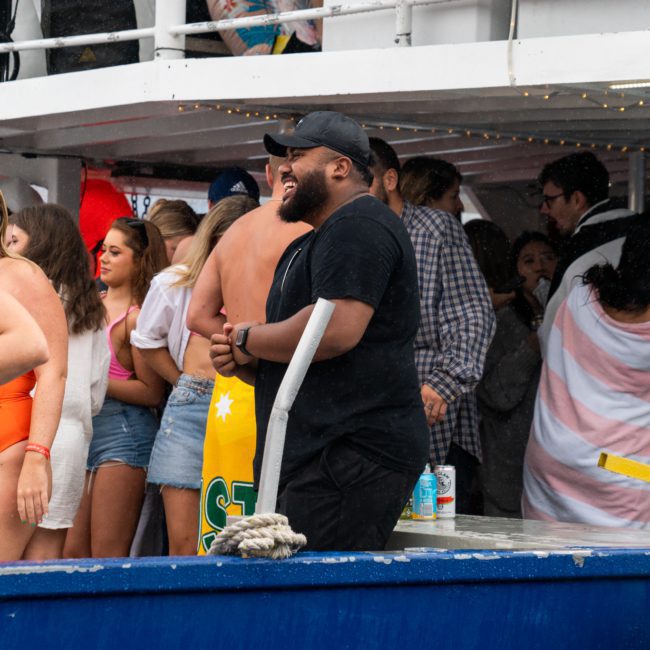 A group of people, some wearing swimsuits, are gathered on a boat. One person in a black shirt stands smiling near the railing, while others socialize under a covered area during a lively Sydney boat party hire.