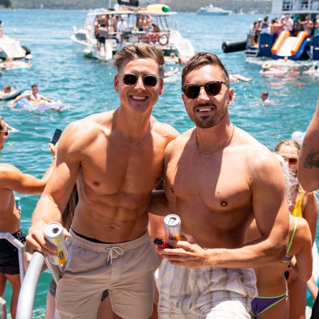 Two men in swim trunks and sunglasses pose with drinks on a luxury yacht hire in Sydney. People swim and relax on nearby boats in the background.