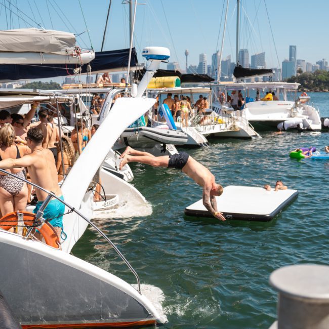 People enjoying a sunny day on various boats and floating devices in a lively water party scene, with one person diving into the water. A catamaran party in Sydney adds an extra touch of excitement, with the city skyline visible in the background.