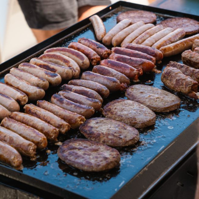 Sausages and patties are cooking on a large outdoor grill with people standing nearby, enjoying a vibrant Sydney boat party hire.