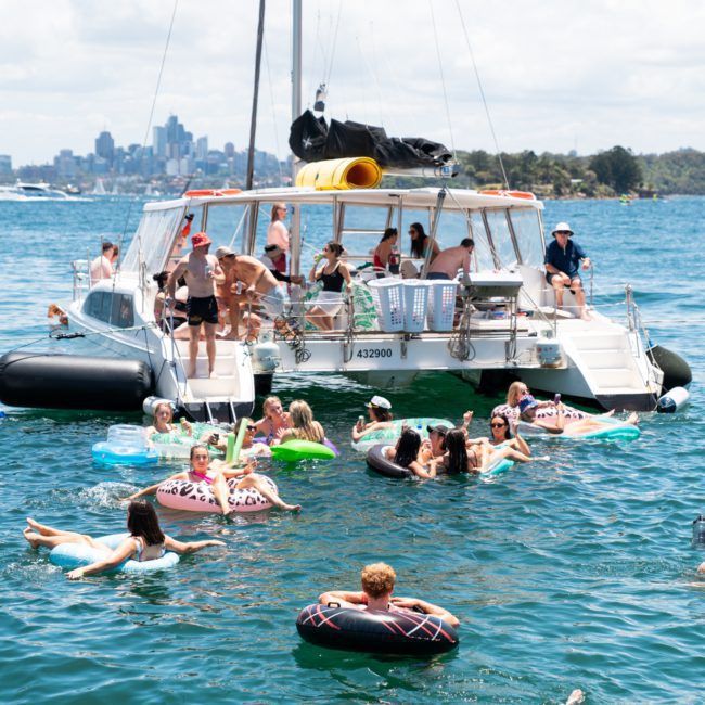A group of people enjoy a sunny day on a catamaran party in Sydney, floating on inflatables and swimming near the boat. The city skyline and other boats are visible in the background.