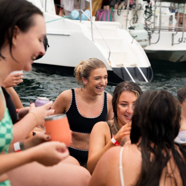 A group of people in swimsuits enjoys a social gathering on the water near a luxury yacht hire Sydney. Some hold drinks as they smile and interact with each other.