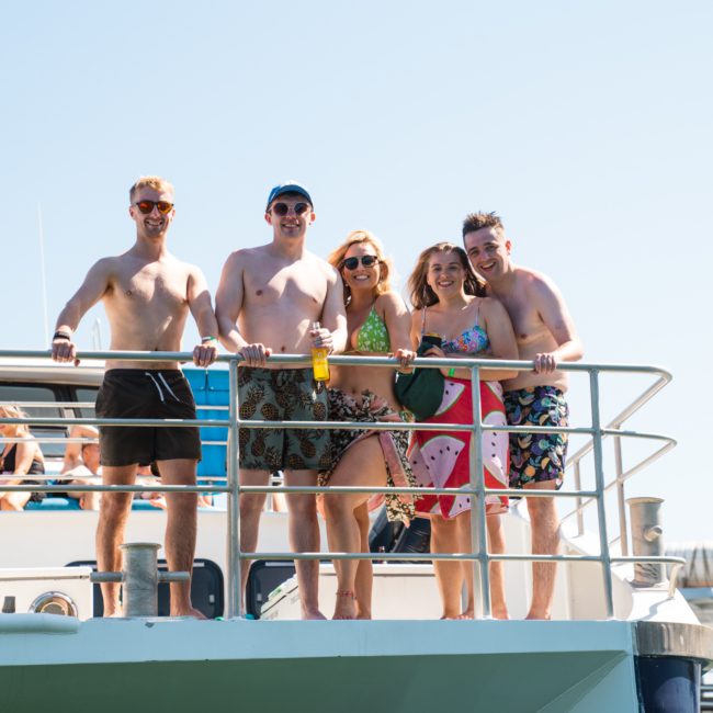 A group of five people stands on the deck of a boat, smiling at the camera. They are wearing swimsuits and summer attire under a clear, sunny sky. It's a lively atmosphere perfect for corporate boat events in Sydney, and other people can be seen enjoying themselves in the background.