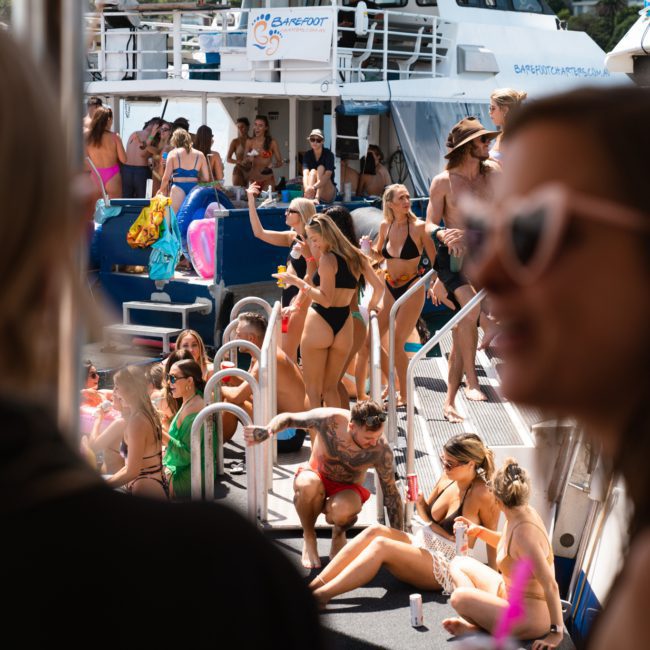 A group of people in swimsuits are socializing and enjoying a sunny day on a catamaran party Sydney. The boat is bustling with activity, some standing while others sit or lean on the railing.