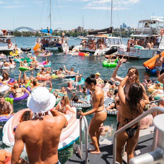 A lively boat party on the water with many people on inflatables and boats, dancing and celebrating under a sunny sky. Several boats and the Sydney Harbour Bridge are visible in the background, adding to the festive atmosphere. Perfect for those considering a Catamaran party Sydney experience!