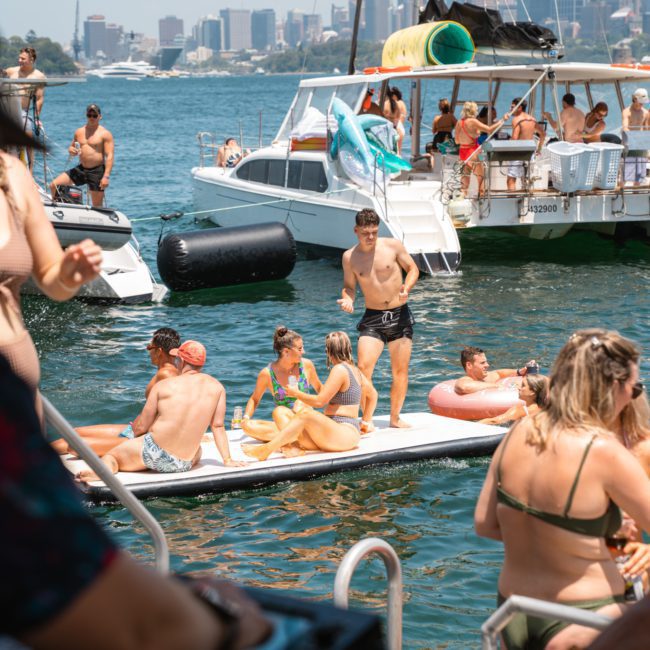 A group of people enjoying a sunny day on boats and inflatables in a harbor, with a visible city skyline in the background. A DJ setup with turntables is in the foreground, creating the perfect atmosphere for a Sydney boat party hire.