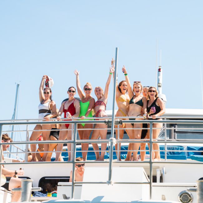 A group of people in swimwear stand on a boat's upper deck, holding drinks and posing for a photo during a vibrant catamaran party in Sydney. Others are on the lower deck, some sitting and one taking a photo, all against the backdrop of a clear blue sky.