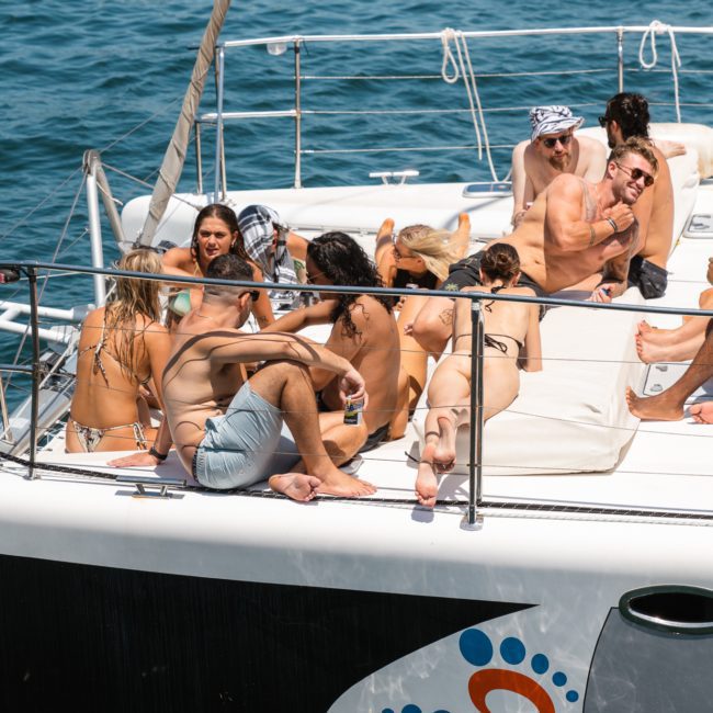 A group of people in swimsuits relax on a private yacht charter Sydney Harbour, with some sitting and others lying on the deck under the midday sun on the water.